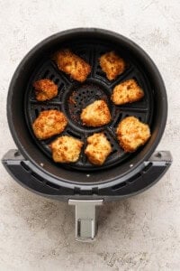 Eight breaded chicken nuggets are arranged in the basket of an air fryer, cooked to a golden brown color on a textured light surface.