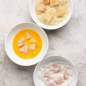 Three white bowls with raw chicken pieces: one with flour, one with beaten egg, and one with breadcrumbs, showing the breading process.