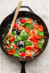 A cast-iron skillet with chopped red and green bell peppers and diced onions being sautéed, stirred with a wooden spoon.