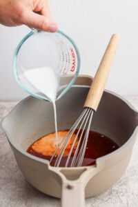 A hand pours a white liquid from a measuring cup into a pot containing a brown sauce, with a whisk resting inside the pot.