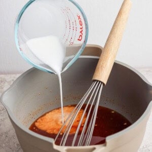 A hand pours a white liquid from a measuring cup into a pot containing a brown sauce, with a whisk resting inside the pot.