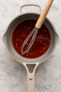 A saucepan containing a reddish-brown sauce with chili flakes, being whisked with a metal whisk on a textured light surface.