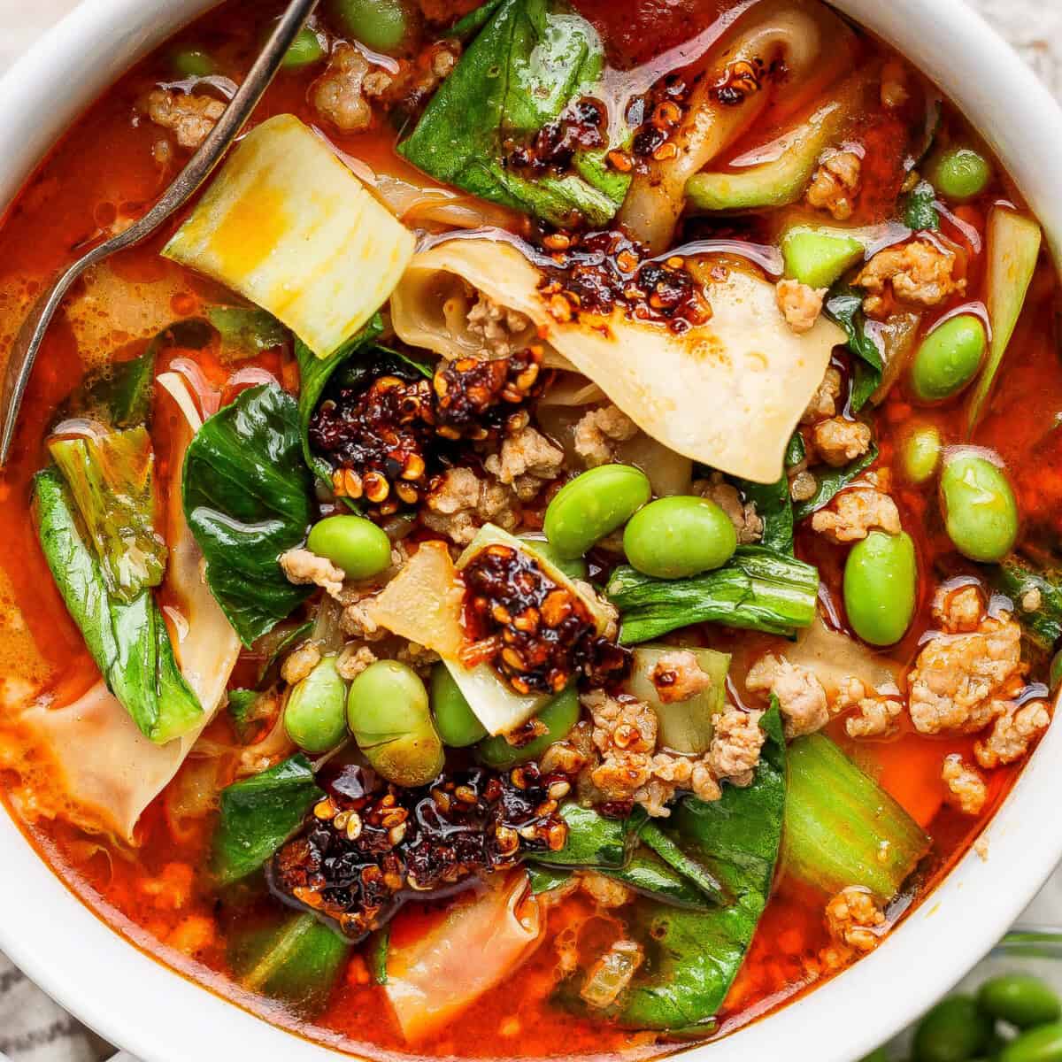 A bowl of spicy noodle soup with ground meat, bok choy, edamame, chili oil, and wide noodles, with a spoon partially visible.