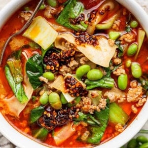 A bowl of spicy noodle soup with ground meat, bok choy, edamame, chili oil, and wide noodles, with a spoon partially visible.