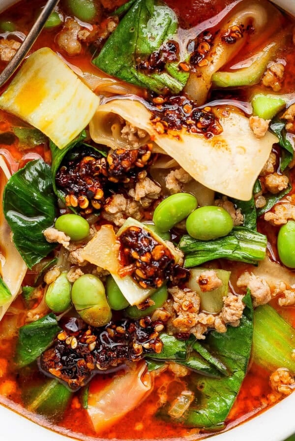 A bowl of spicy noodle soup with ground meat, bok choy, edamame, chili oil, and wide noodles, with a spoon partially visible.