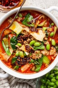 A bowl of spicy noodle soup with ground meat, bok choy, edamame, chili oil, and wide noodles, served with a metal spoon.