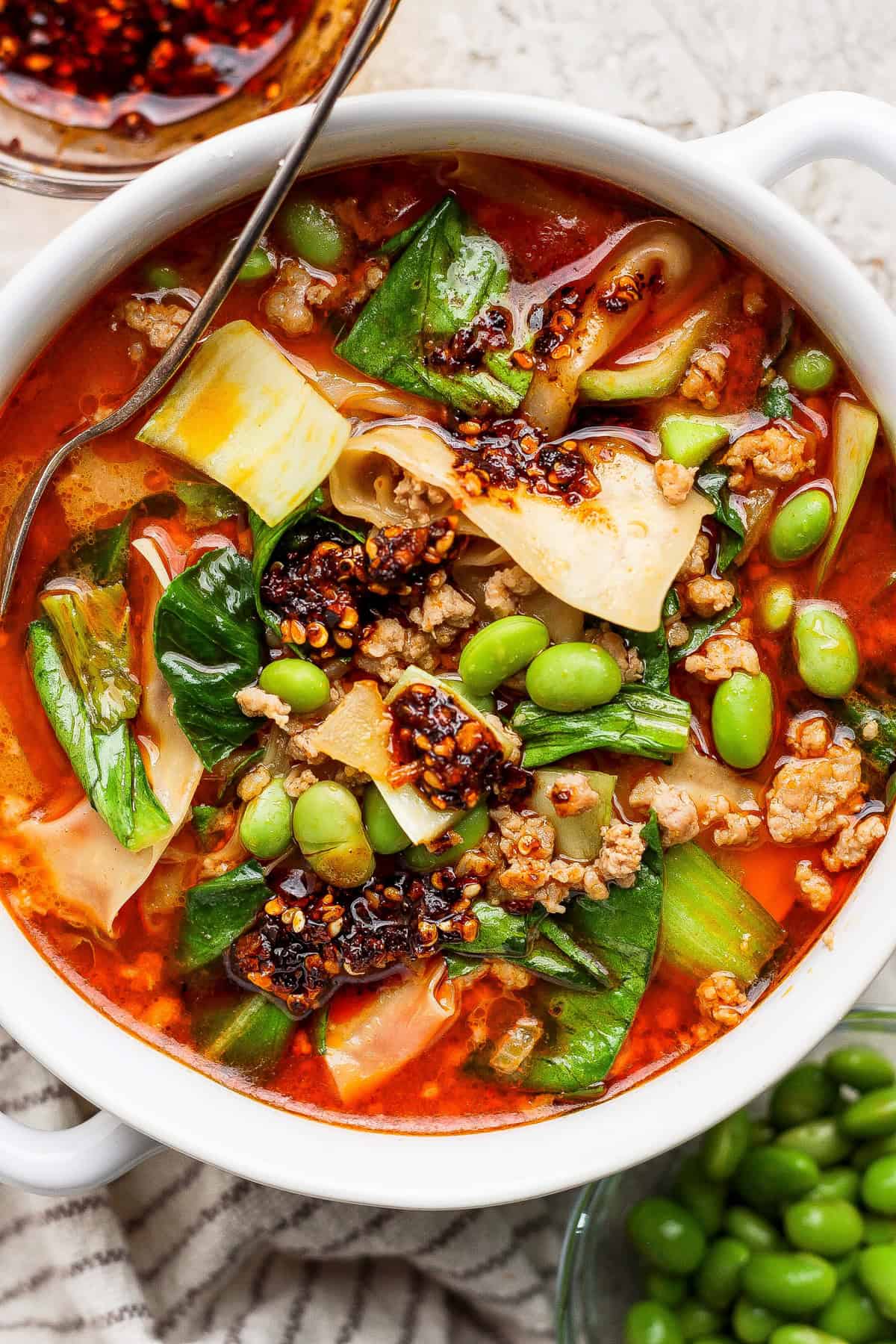 A bowl of spicy noodle soup with ground meat, bok choy, edamame, chili oil, and wide noodles, served with a metal spoon.