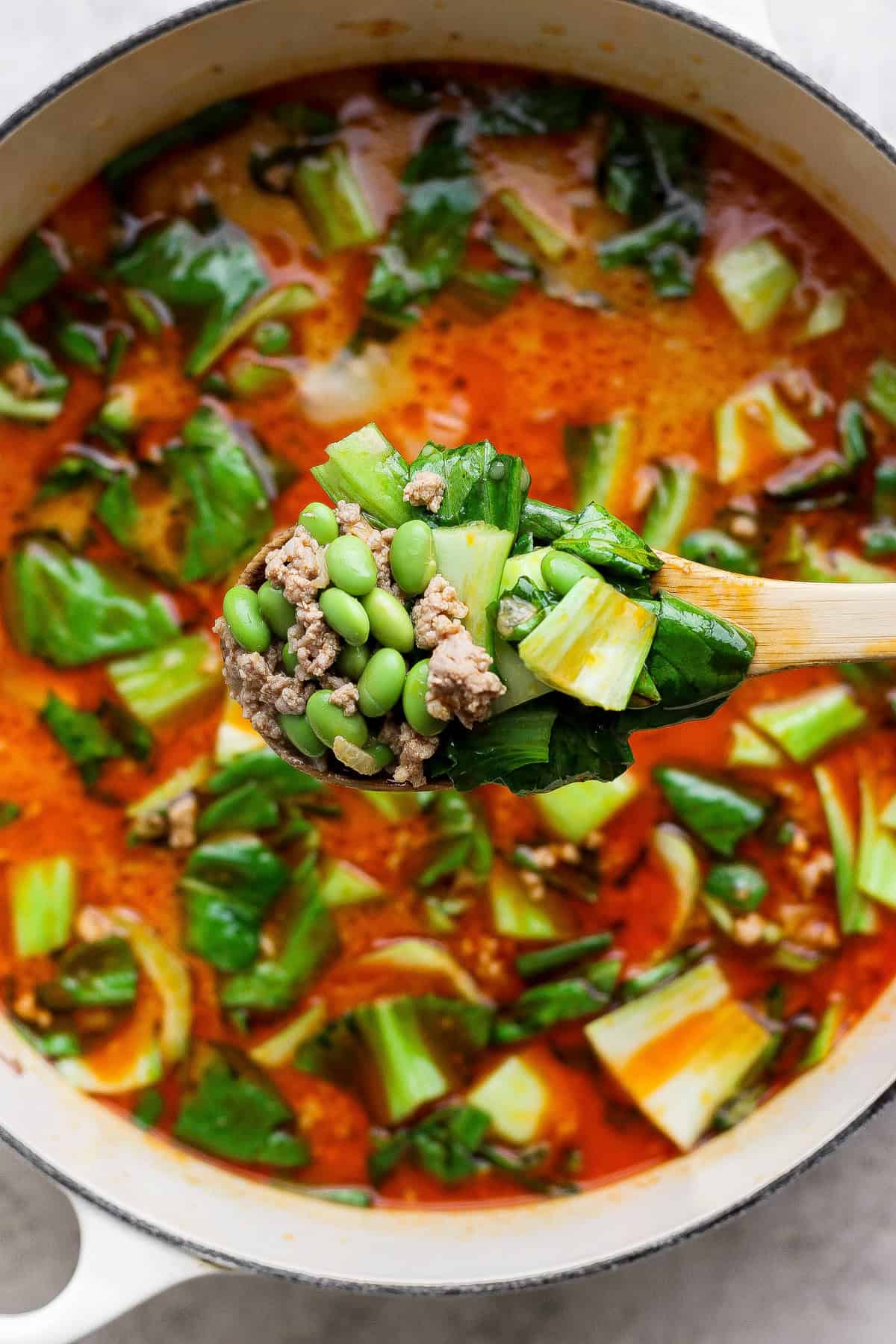A wooden spoon holds a mix of ground meat, edamame, and bok choy over a pot of red broth soup with leafy greens.