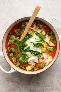 A pot of red broth soup with leafy greens, noodles, and vegetables, stirred with a wooden spoon, set on a light textured surface.
