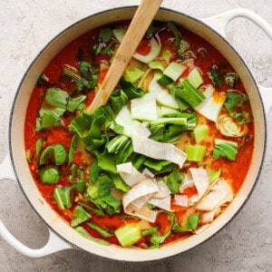 A pot of red broth soup with leafy greens, noodles, and vegetables, stirred with a wooden spoon, set on a light textured surface.