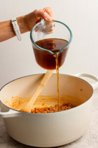 A hand pours broth from a glass measuring cup into a white pot containing ground meat and a wooden spoon.