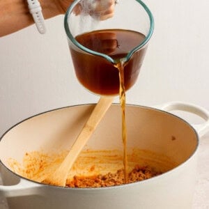 A hand pours broth from a glass measuring cup into a white pot containing ground meat and a wooden spoon.