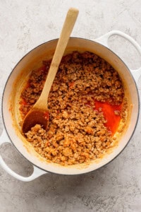Cooked ground meat with tomato sauce in a white pot, stirred with a wooden spoon, on a light textured surface.