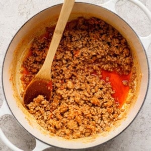 Cooked ground meat with tomato sauce in a white pot, stirred with a wooden spoon, on a light textured surface.