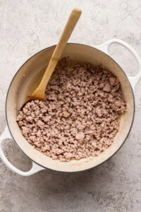Cooked ground meat in a white pot with a wooden spoon, photographed from above on a light textured surface.