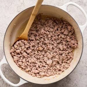 Cooked ground meat in a white pot with a wooden spoon, photographed from above on a light textured surface.