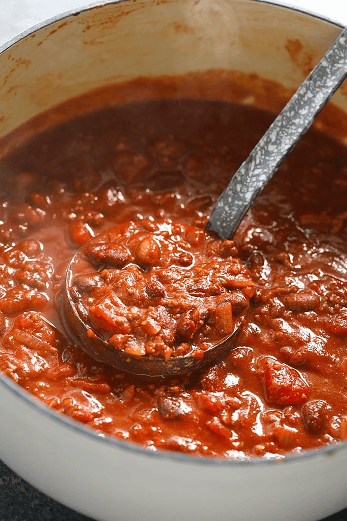 A ladle scoops thick chili with beans, tomato chunks, and ground meat from a large white pot—an inviting chili recipe in progress.