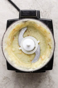 Top view of a food processor containing a pale, creamy blended mixture with visible metal blades in the center.