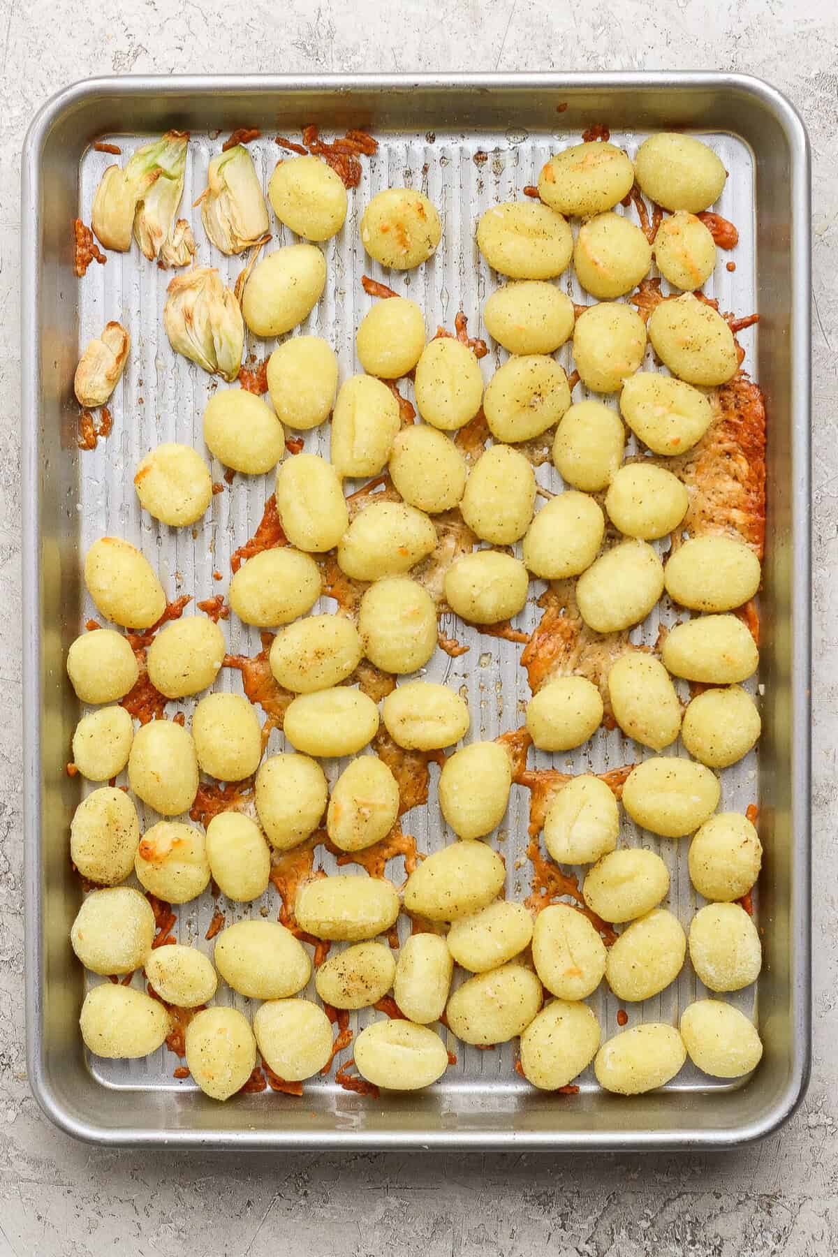 A metal baking sheet with scattered cooked gnocchi, some clumps of melted cheese, and a few roasted garlic cloves on a textured surface.