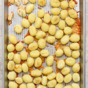 A metal baking sheet with scattered cooked gnocchi, some clumps of melted cheese, and a few roasted garlic cloves on a textured surface.
