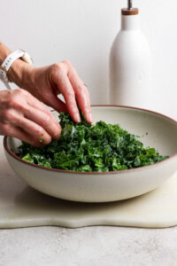 A person massages chopped kale in a large bowl, with a white bottle in the background on a light-colored surface.