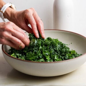 A person massages chopped kale in a large bowl, with a white bottle in the background on a light-colored surface.