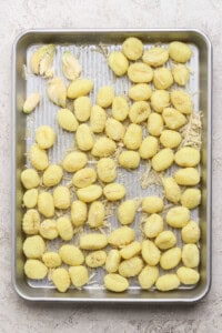A metal baking tray with uncooked gnocchi, shredded cheese, and garlic cloves arranged on a light, textured surface.