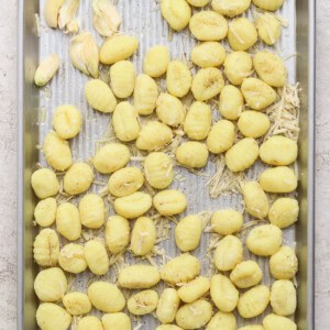 A metal baking tray with uncooked gnocchi, shredded cheese, and garlic cloves arranged on a light, textured surface.