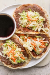 Three tacos filled with refried beans, shredded cabbage, and carrots are served on a white plate with a small bowl of dark dipping sauce.