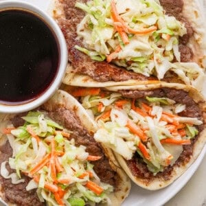 Three tacos filled with refried beans, shredded cabbage, and carrots are served on a white plate with a small bowl of dark dipping sauce.