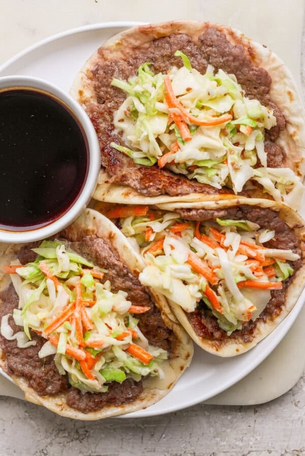 Three tacos filled with refried beans, shredded cabbage, and carrots are served on a white plate with a small bowl of dark dipping sauce.