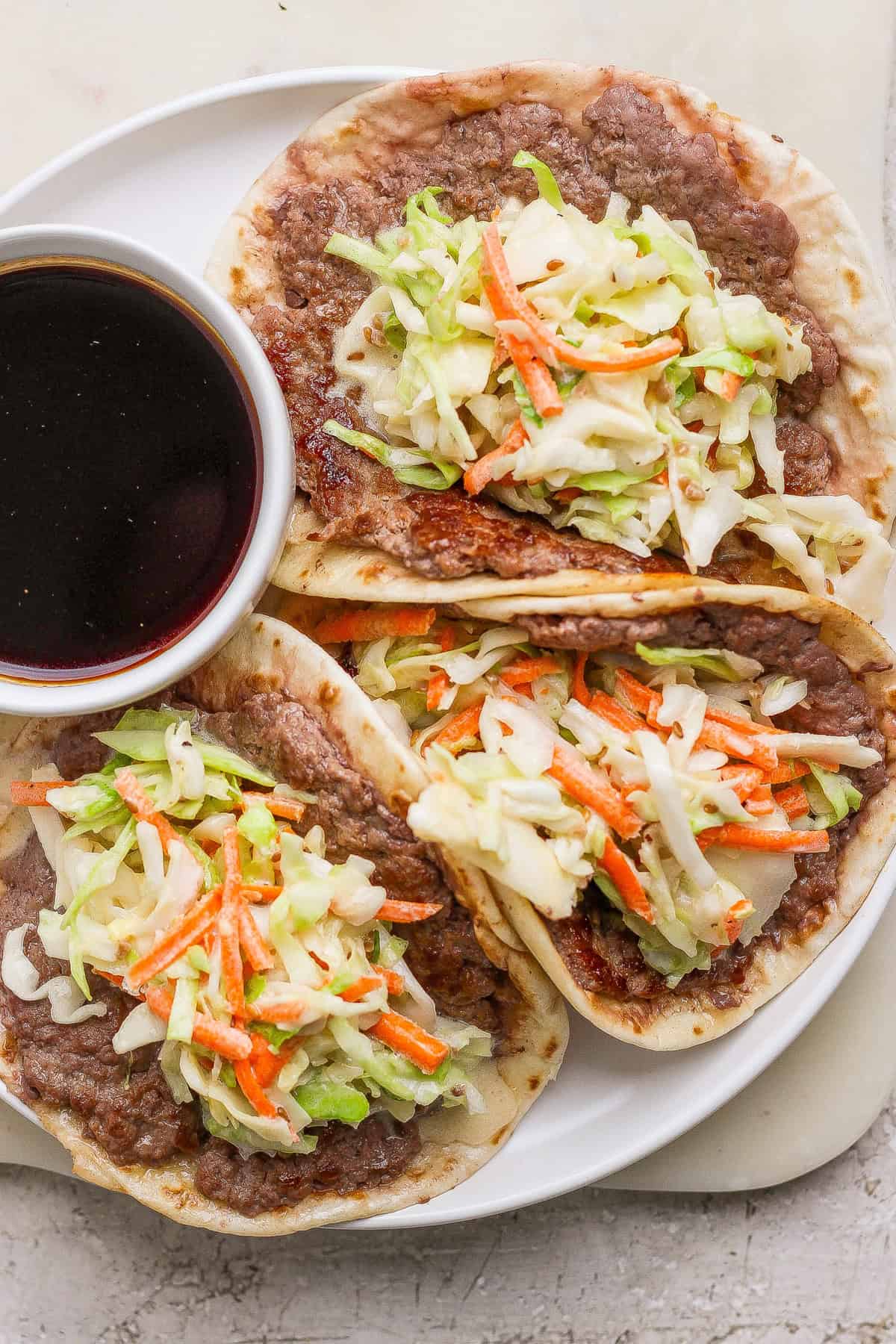 Three tacos filled with refried beans, shredded cabbage, and carrots are served on a white plate with a small bowl of dark dipping sauce.
