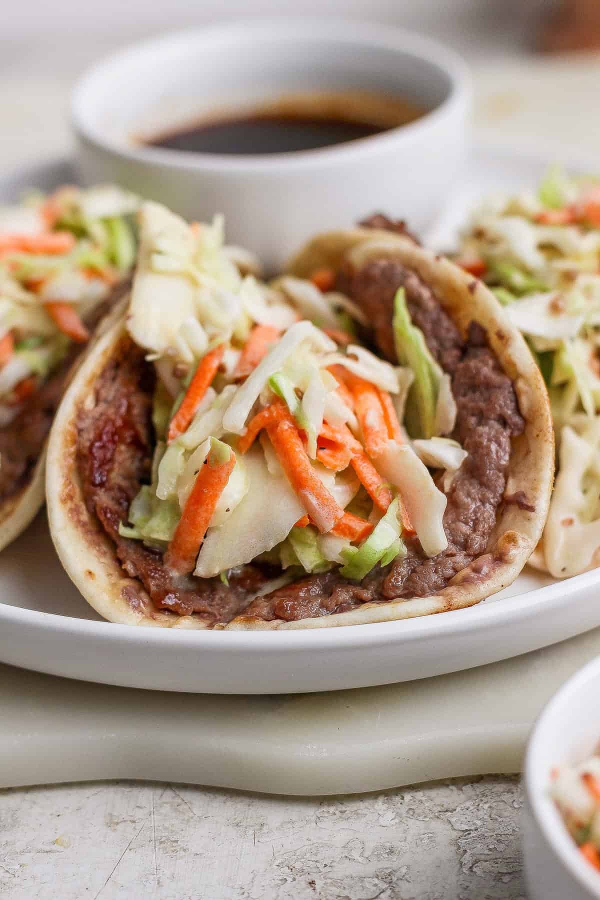 A soft taco filled with refried beans and topped with shredded cabbage and carrots is served on a white plate with a small bowl of sauce in the background.
