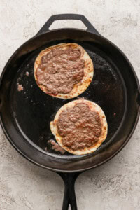Two corn tortillas topped with ground meat cooking in a black cast iron skillet on a light-colored textured surface.