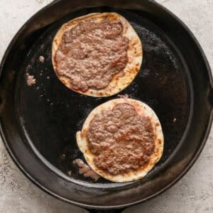 Two corn tortillas topped with ground meat cooking in a black cast iron skillet on a light-colored textured surface.