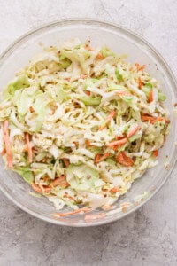 A glass bowl filled with coleslaw made of shredded cabbage and carrots on a light, textured surface.