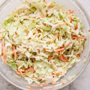 A glass bowl filled with coleslaw made of shredded cabbage and carrots on a light, textured surface.