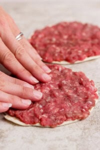 Two hands press raw ground meat onto a flatbread or dough circle, forming a thin layer. Another prepared flatbread with meat is visible in the background.