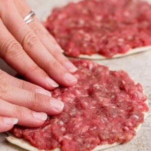 Two hands press raw ground meat onto a flatbread or dough circle, forming a thin layer. Another prepared flatbread with meat is visible in the background.