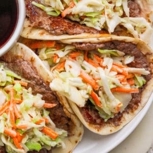Three tacos filled with cooked beef and topped with shredded cabbage and carrots, served on a plate next to a small bowl of dark sauce.
