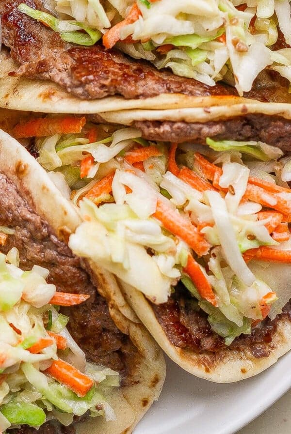 Three tacos filled with cooked beef and topped with shredded cabbage and carrots, served on a plate next to a small bowl of dark sauce.