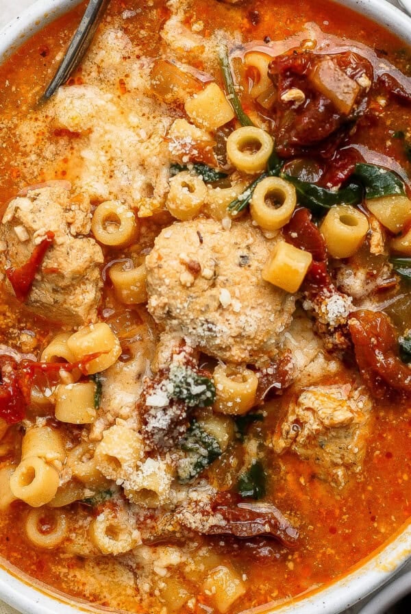 A bowl of tomato-based soup with meatballs, ditalini pasta, sun-dried tomatoes, spinach, and grated cheese on top.