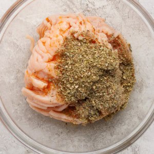 Raw ground meat topped with dried herbs and spices in a clear glass bowl on a light textured surface.