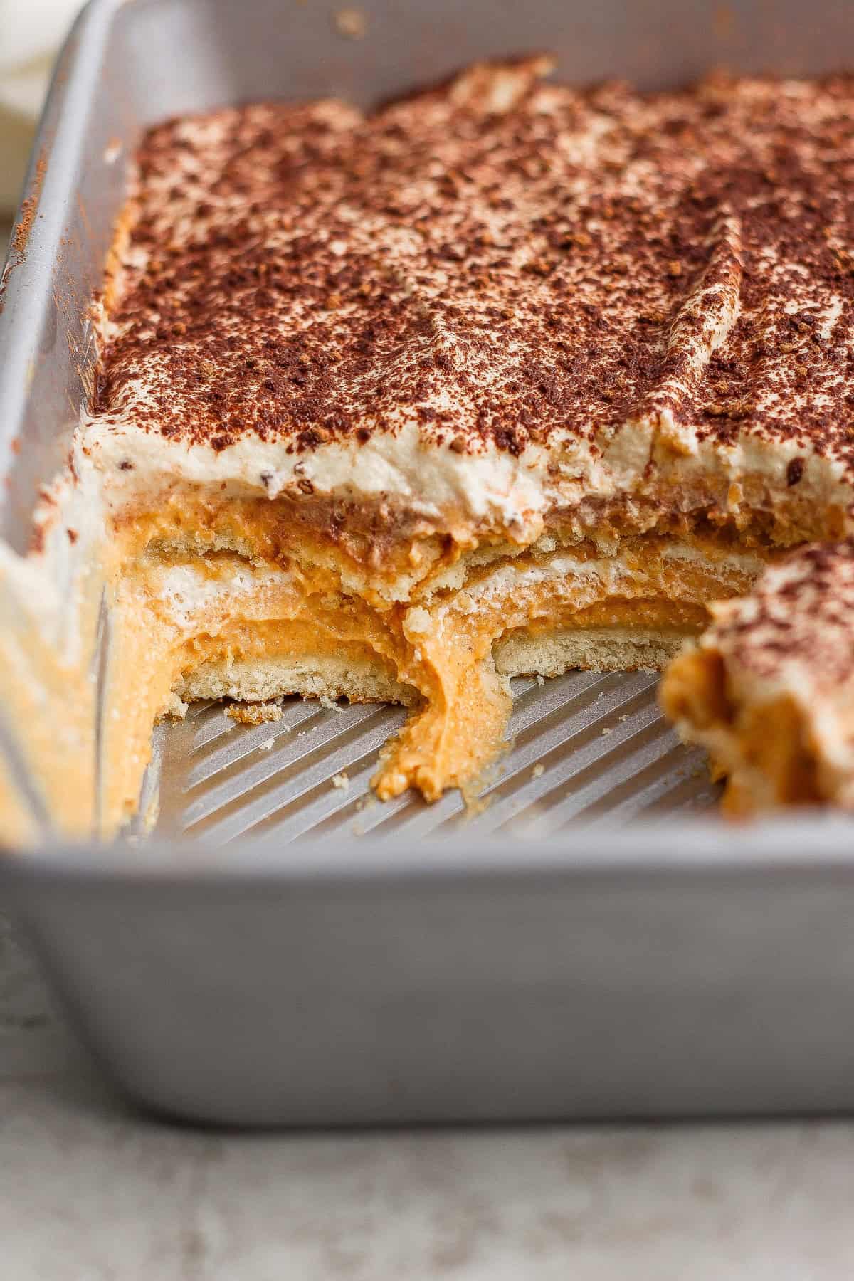 A rectangular baking pan containing a layered dessert with a creamy filling and a dusting of cocoa powder, with a serving removed to show the layers.