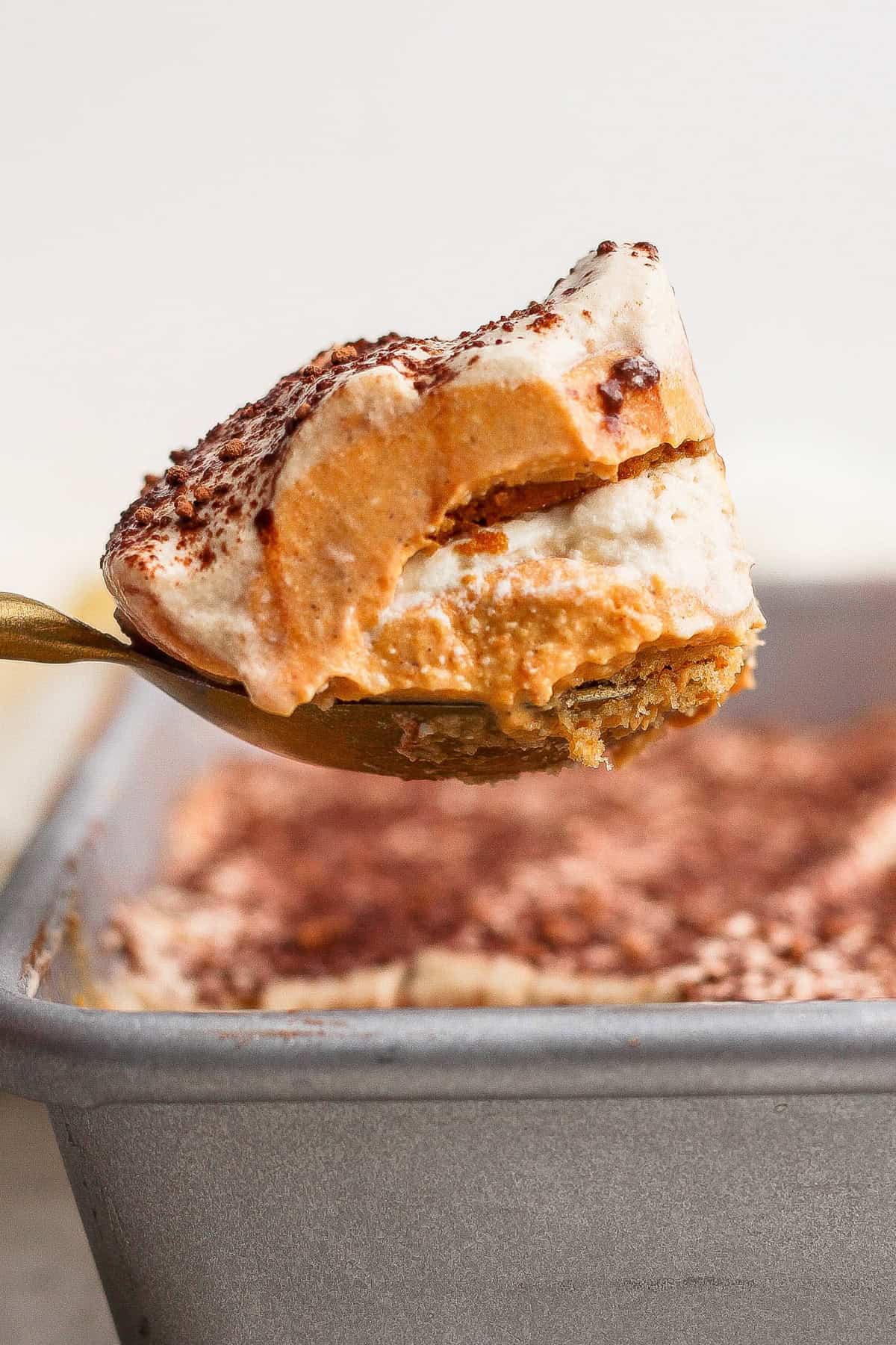 A spoon lifts a layered dessert with a creamy top dusted with cocoa powder from a baking pan.