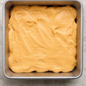 A square metal baking pan filled with an even layer of light brown cake batter on a textured light surface.