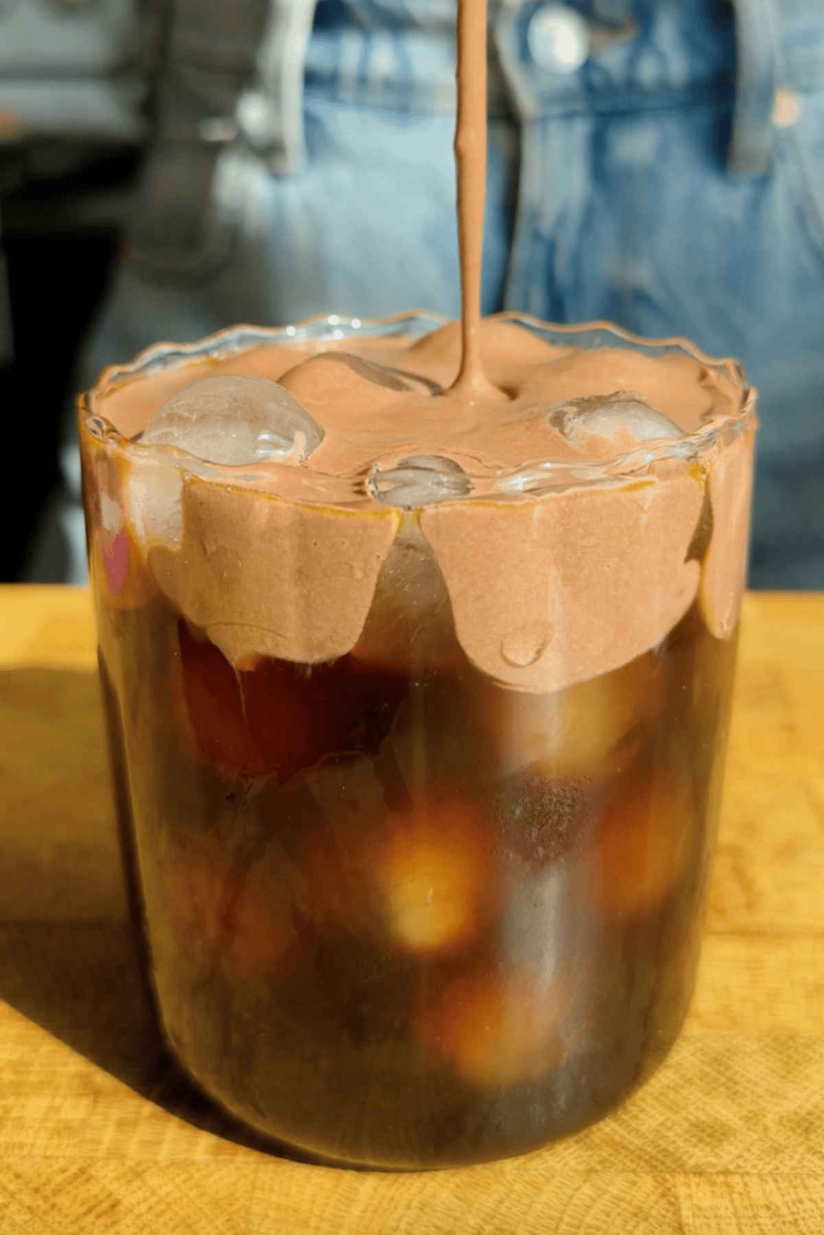 A glass of iced coffee topped with whipped chocolate cream, with someone pouring more cream on top. Denim clothing is visible in the background.