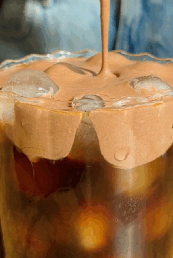 A glass of iced coffee topped with whipped chocolate foam, with the foam being poured over the ice. Denim fabric is visible in the background.