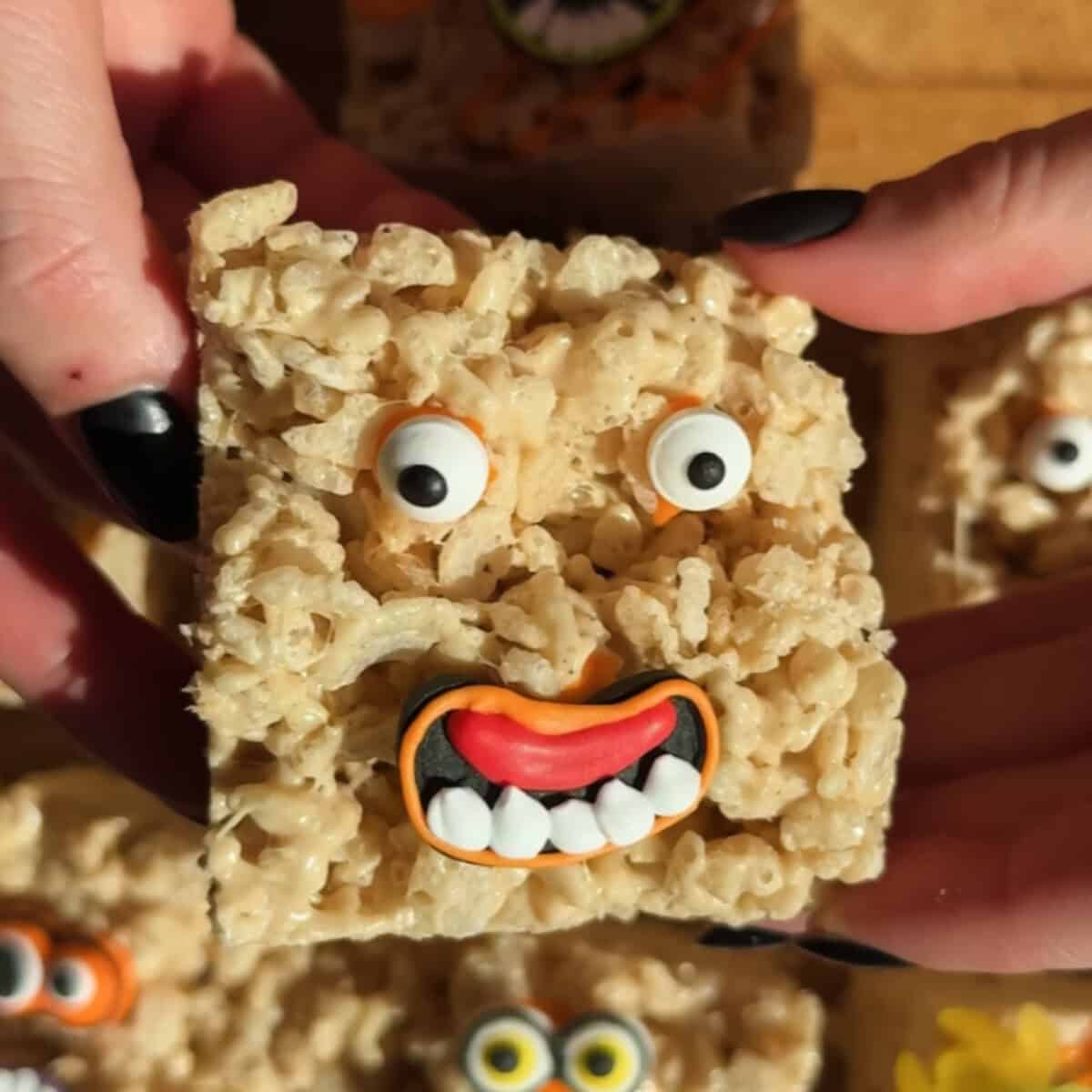 A Rice Krispies treat decorated with candy eyes and a mouth, resembling a goofy face, is held between two hands with black-painted nails.