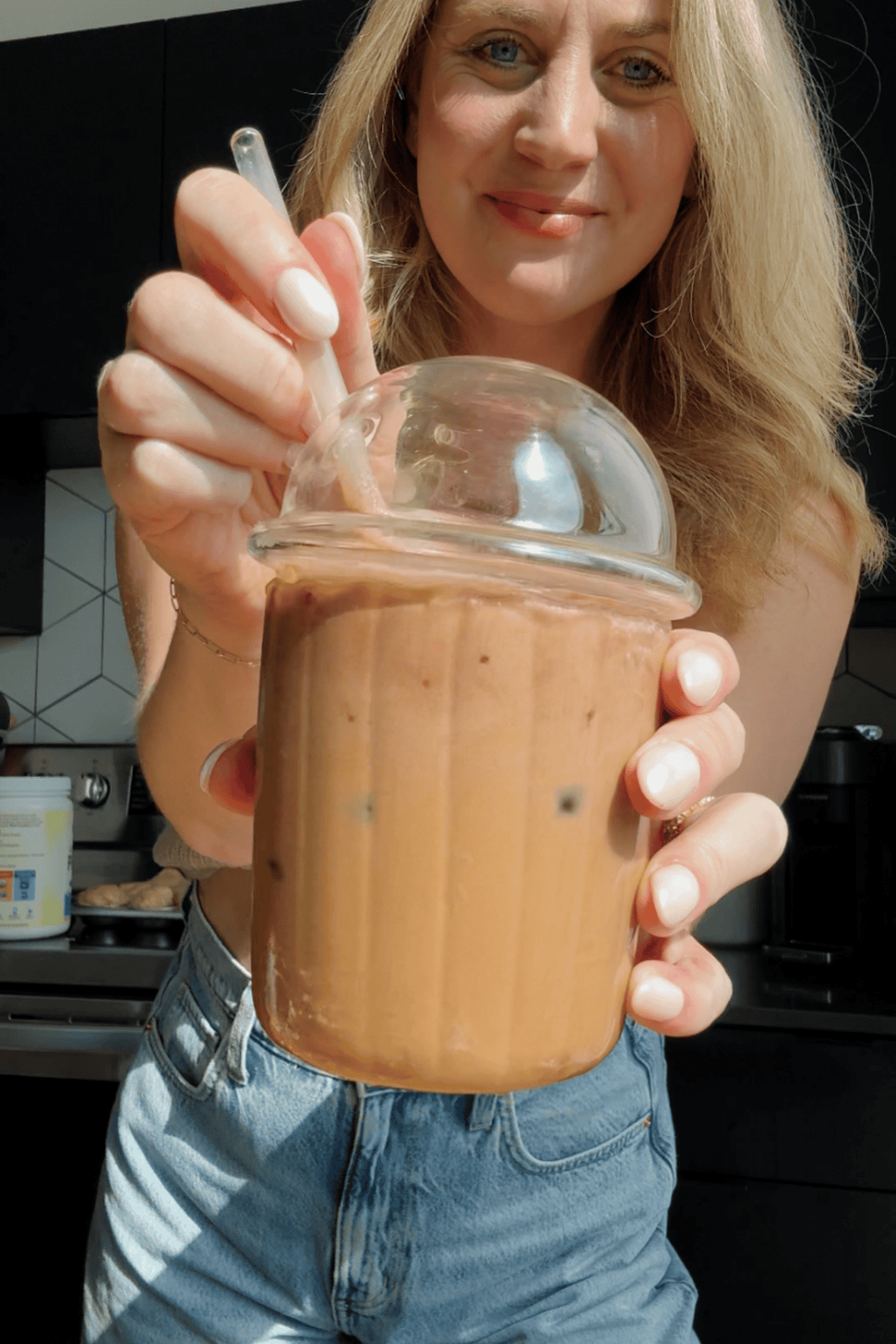 A woman holds a plastic cup of iced coffee with a dome lid and straw, topped with protein cold foam, standing in a kitchen.